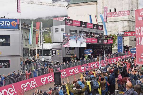 Egan Bernal rua in piaza, in Anpezo, ai 24 de maśo del 2021, sote el cianpanin de San Iaco, vestì de rośa.
